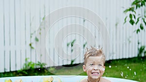 A four-year-old boy lies in a children`s inflatable pool, splashing water, smiling. Nearby float toys. In the garden, in