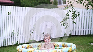 A four-year-old boy lies in a children`s inflatable pool, splashing water, smiling. Nearby float toys. In the garden, in