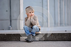 A four-year-old white blonde boy on a gray concrete background sitting on on the steps, pensive photo