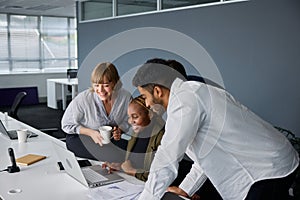 Four young adults in businesswear smiling while working on laptop at desk in office