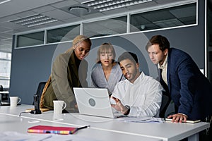 Four young adults in businesswear talking and gesturing by laptop on desk in office