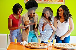 Four young african girls in bright colored fast food restaurant making photo of pizza in their mobile phones