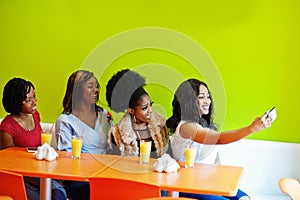 Four young african girls with juices sitting in bright colored fast food restaurant and making selfie by mobile phone