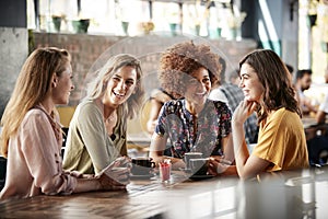 Four Young Female Friends Meeting Sit At Table In Coffee Shop And Talk