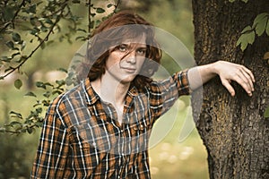 Foxy man with long carroty hair stands leaning on a tree trunk. Young handsome freckled guy in chequered shirt looking