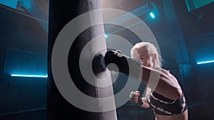 Fragile young woman delivering powerful punches to punchbag. Female athlete attacking punching bag in dark gym with blue