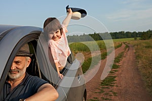 A fragment of a car on a field road with an adult man driving and a happy little girl in the back window.