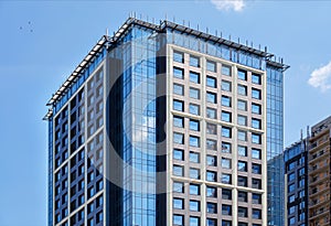 A fragment of the glass facade of a modern concrete building under construction with a reflection of the blue sky and a tower