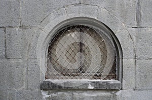 fragment of a historic stone building with a small arched window frame. Old arch window with grille. Bars on the windowframe. grey