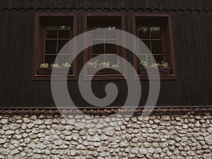 Fragment of a wall of an old brown wooden house with three windows and a stone basement.