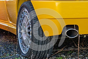 Fragment of yellow sport car with rear left weel parked on gravel road .