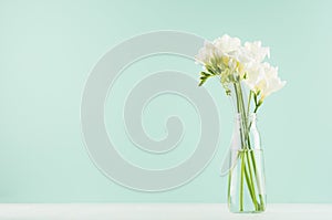 Fragrance soft light white flowers freesia in bouquet in simple glass bottle in green mint menthe interior on white wood board.