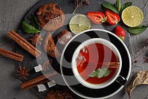 Fragrant tea in a black cup on a black plate with biscuits, lemon, cinnamon and fruits
