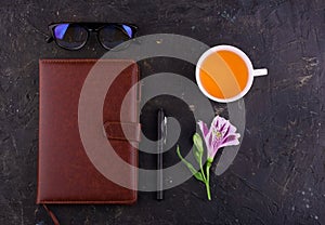 Fragrant tea in a white cup, beautiful flowers, a black background, a notebook with a pen and glasses