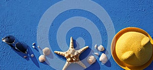 Frame with beach accessories on a nautical theme: yellow straw hat, sunglasses, starfish and shells on a blue background.