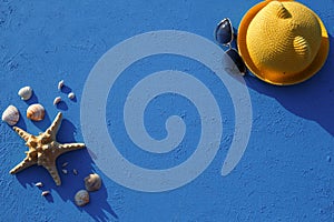 Frame with beach accessories on a nautical theme: yellow straw hat, sunglasses, starfish and shells on a blue background.