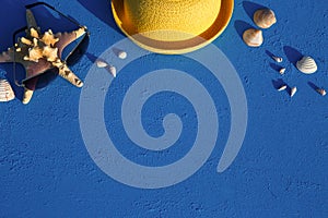 Frame with beach accessories on a nautical theme: yellow straw hat, sunglasses, starfish and shells on a blue background.