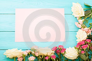 Frame of colorful roses on blue wooden background with paper card and copy space. Top view and selective focus. Mock up