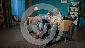 In the frame, an elderly couple sits on a couch in an apartment, against a blue wall. They are looking at, leafing