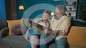 In the frame, an elderly couple sits on a couch in an apartment, against a blue wall. They are looking at a photo album