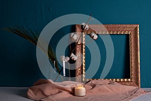 Frame for a picture, a candle is burning nearby and there is a vase with a dry plant of cotton and fern on a blue background