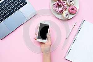 Frame woman`s hands holding white smartphone with black copyspace on pink background with other accessories. Feminine business mo