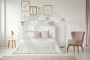 Framed posters gallery above a cozy double bed in a feminine room interior with a comfy, retro armchair