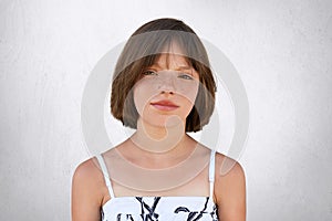 Freckled girl with hazel eyes and dark short hair, looking with displeasure into camera while posing against whiye concrete wall. photo