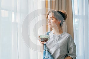 Free Time To Relax. Young woman looking out the window while overlooking the city at cozy home holding cup of hot tea