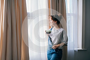 Free Time To Relax. Young woman looking out the window while overlooking the city at cozy home holding cup of hot tea