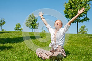 Freedom and happiness concept. Beautiful woman with raising up her hands is sitting on grass, enjoy sunny day