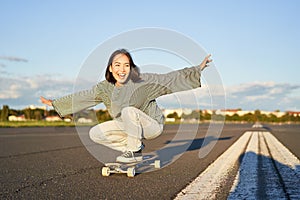 Freedom and happiness. Happy asian girl riding her longboard on an empty sunny road, laughing and smiling, skateboarding