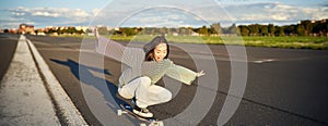 Freedom and happiness. Happy asian girl riding her longboard on an empty sunny road, laughing and smiling, skateboarding