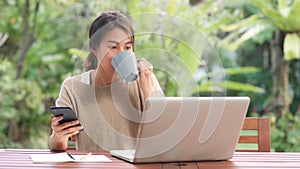 Freelance Asian woman working at home, business female working on laptop and using mobile phone drinking coffee sitting on table