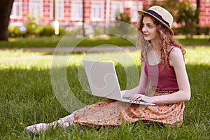 Freelance business concept. Woman sitting on green grass lawn in city park, working on laptop, has pensive facial expression,