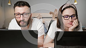 Freelance family working at home concept. Man and woman lying on bed in bedroom and simultaneously working typing on