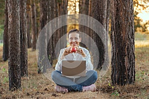 A freelance girl is sitting with a laptop on her lap and holding a ripe red apple in a pine forest