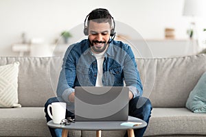 Freelance job. Young smiling Arab man working on laptop from home, wearing headphones, using pc computer