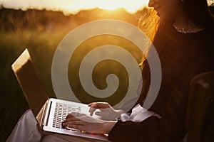 Freelance and remote work outdoors. Fashionable elegant girl with hands on keyboard sitting in sunny summer field at sunset.Young