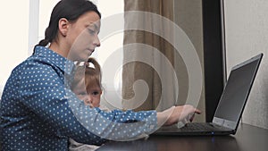 A freelance woman with a small child in her arms works at a laptop in her home office, a girl plays with cubes while
