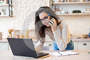 Freelance woman working from home, sitting at kitchen table, using laptop and having phone conversation