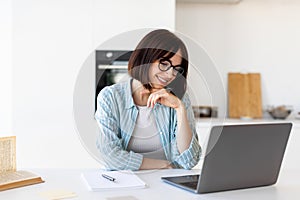 Freelance work concept. Happy young lady sitting at table with laptop in kitchen, looking at screen and smiling