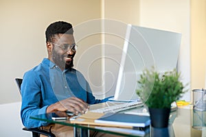 Freelance work. Happy black man working on computer at workpalce, looking at screen and smiling, enjoying remote job