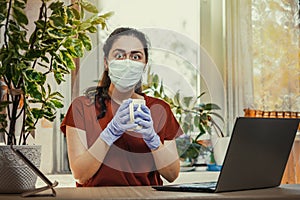 Freelance.A young Caucasian woman in gloves and a medical mask, holding a Cup of tea, and her eyes bulged. Home office