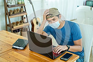 Freelance young man wearing face mask working with laptop computer and feeling sad about failing to invest online in the stock