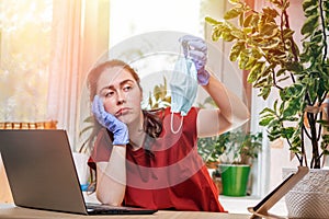 Freelance.A young pretty woman in rubber gloves holds a medical mask with a sad face. Home decor, the sun outside the window.The