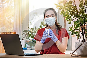 Freelance.A young woman in gloves and a medical mask, holding a Cup of tea, and her eyes bulged. Home office, laptop on the table.