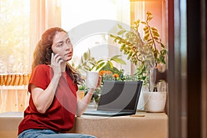 Freelance.A young woman is talking on the phone, and holding a mug in her hand.Home decor,the sun outside the window and a laptop