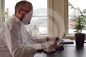 Freelancer bearded man 50 years old in a white shirt sits at a table and takes notes in a laptop.