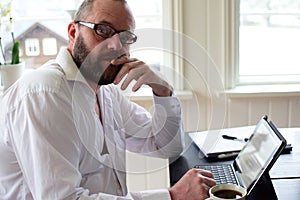 Freelancer bearded man 50 years old in a white shirt sits at a table and takes notes in a laptop.